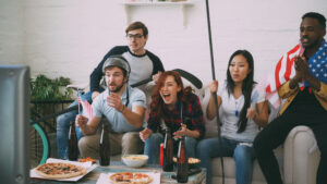 Multi ethnic group of friends sports fans with USA national flags watching winter olympic games on TV together cheering up favorite team at home indoors